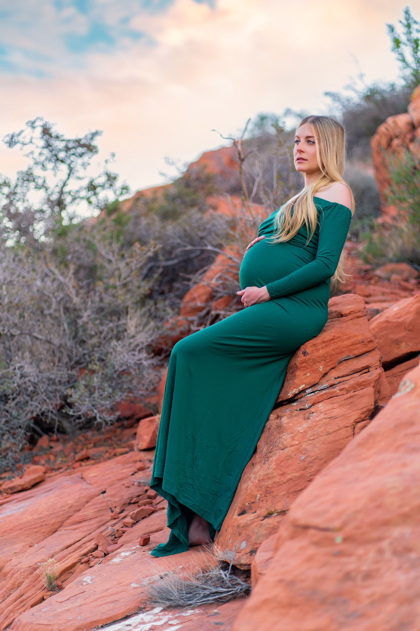 Expectant mother in an emerald green gown resting on red rock formations at dusk